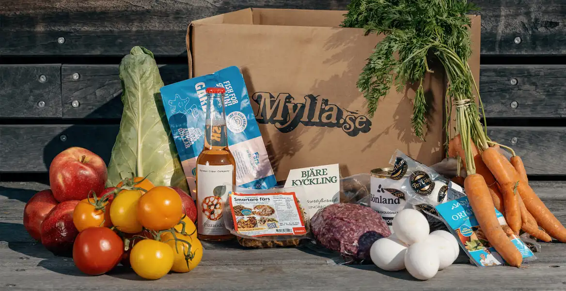 En papperslåda med texten "Mylla.se" står på en träyta utomhus, omgiven av färska matvaror. Framför lådan syns en ciderflaska, en förpackning kyckling, köttfärs, ekologiska ägg, gula och röda tomater, samt en förpackning med fisk. Grönsaker och rotfrukter kompletterar bilden, och bakgrunden består av en rustik trävägg.