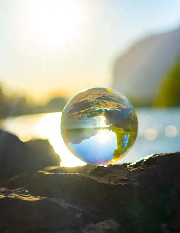 En genomskinlig glaskula placerad på en klippa vid vattnet, som reflekterar och inverterar landskapet med himmel, moln och solnedgång, medan bakgrunden är oskarp med varma ljusreflektioner på vattnet.